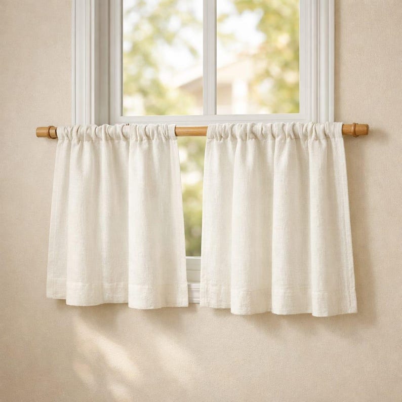 Puede incluir: Un par de cortinas de caf&eacute; de color blanco roto hechas de una tela texturizada, fruncidas en una barra de madera de color marr&oacute;n claro. Las cortinas cuelgan en pliegues verticales, cubriendo la parte inferior del marco de la ventana.