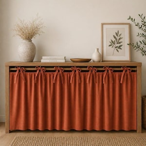 Puede incluir: Un mueble de madera con una cortina de color terracota asegurada con lazos. El mueble está coronado con objetos decorativos, incluyendo una impresión botánica enmarcada, un jarrón y libros. La estética general es cálida y acogedora.