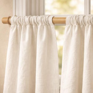 Puede incluir: Un par de cortinas de tela texturizada en color blanco roto colgadas de una barra de madera clara. Las cortinas presentan un bolsillo superior fruncido, creando pliegues verticales que caen hacia abajo, cubriendo parcialmente el marco.