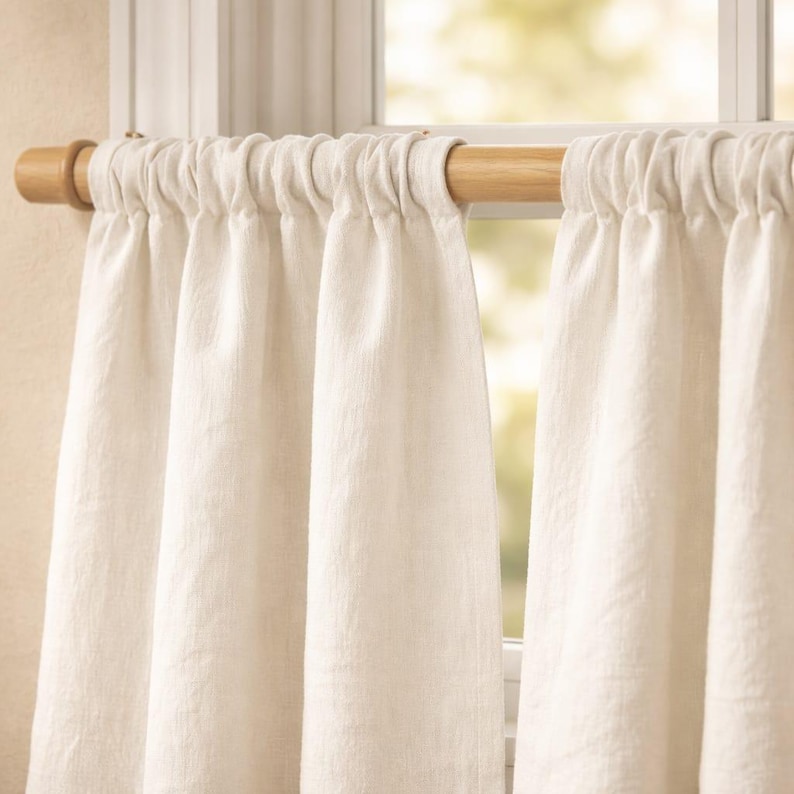 Puede incluir: Un par de cortinas de tela texturizada en color blanco roto colgadas de una barra de madera clara. Las cortinas presentan un bolsillo superior fruncido, creando pliegues verticales que caen hacia abajo, cubriendo parcialmente el marco.