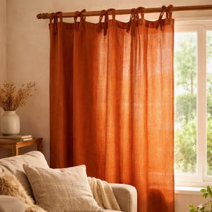May include: Burnt orange linen curtains suspended from a wooden pole with tie fastenings. The curtains, a solid colour, partially obscure a window. A sofa with cushions and a vase of dried flowers are in the foreground.