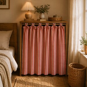 May include: A wooden cabinet with red and white striped curtains. The curtains are tied at the top with bows. A lamp, vase, and basket sit on top of the cabinet. A bed and window are visible in the background. The room has a warm, inviting atmosphere.