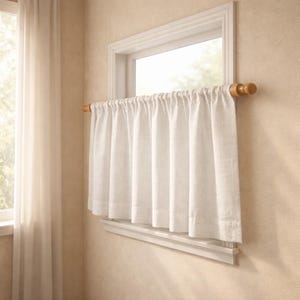 Puede incluir: Una cortina de caf&eacute; blanca cuelga de una barra de madera montada dentro del marco de una ventana. La cortina presenta un encabezado de bolsillo para barra simple y un dobladillo recto, con pliegues verticales suaves y fruncidos.