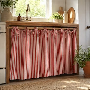 May include: Red and white striped kitchen curtain, tied with bows, hangs below a wooden countertop. The curtain is made of fabric with vertical stripes. The kitchen has a window, a sink, and a neutral-colored rug.
