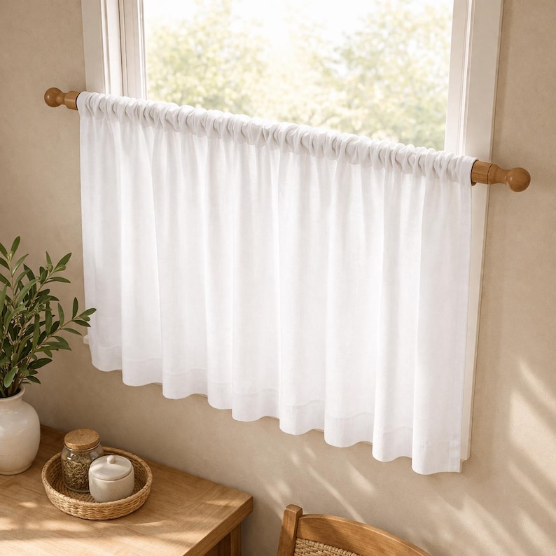 Puede incluir: Una cortina de caf&eacute; blanca y lisa cuelga de una barra de madera con remates redondeados. La cortina presenta un encabezado fruncido y un dobladillo recto, proporcionando una cobertura ligera sobre el cristal de la ventana.
