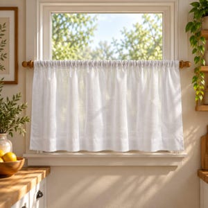 Puede incluir: Una cortina de caf&eacute; blanca cuelga de una barra de madera sobre una ventana. La cortina presenta una parte superior fruncida y un dobladillo ancho en la parte inferior, hecha de una tela semitransparente que permite el paso de la luz.