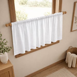Puede incluir: Una cortina de caf&eacute; blanca y lisa cuelga de una barra de madera montada dentro del marco de la ventana. La cortina presenta un encabezado fruncido y un dobladillo recto, proporcionando una ligera cobertura en la parte inferior.