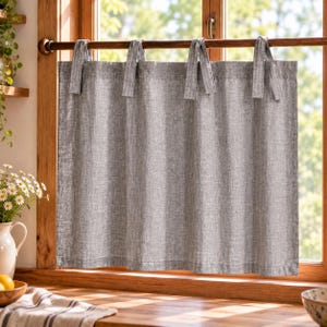 May include: A grey, textured fabric cafe curtain hangs from a dark wooden rod using four fabric tie-top loops. The curtain features a woven, linen-like appearance and covers the lower portion of a window, allowing light to filter through.
