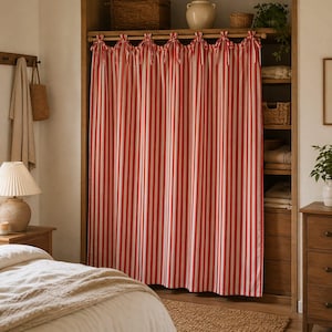 May include: Red and white striped curtain hanging in a wooden closet. The curtain is tied at the top with matching red ribbons. The closet has shelves with baskets, a vase, and plants. A bed and nightstand are in the foreground.