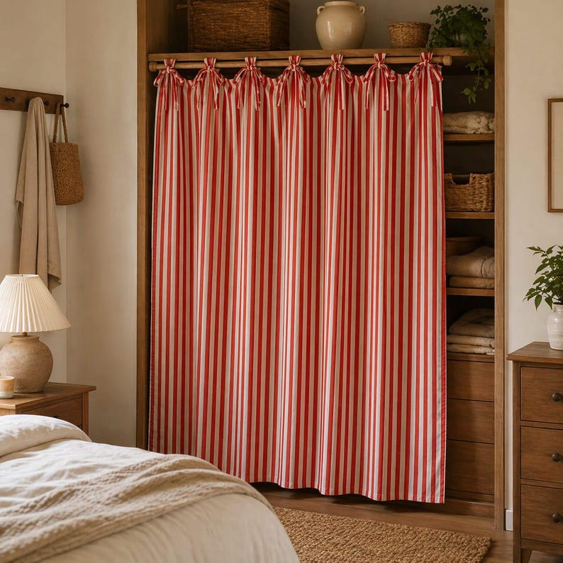 May include: Red and white striped curtain hanging in a wooden closet. The curtain is tied at the top with matching red ribbons. The closet has shelves with baskets, a vase, and plants. A bed and nightstand are in the foreground.
