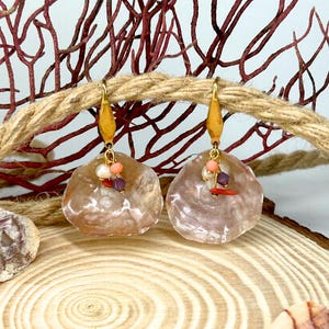 Puede incluir: Pendientes de concha con ganchos dorados. Cada pendiente presenta una concha grande e iridiscente, adornada con coral, perlas y cuentas moradas. Los pendientes se exhiben sobre una superficie de madera, con una cuerda y ramas de coral en el fondo.
