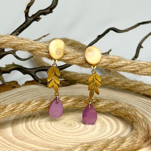 Puede incluir: Un par de pendientes colgantes con una parte superior en espiral color crema, un diseño de hojas doradas y una parte inferior púrpura en forma de lágrima. Los pendientes se exhiben sobre un fondo de cuerda y madera natural.