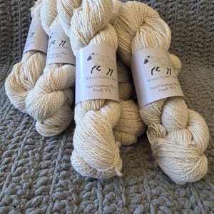 May include: Several skeins of off-white yarn are arranged on a gray, textured surface. Each skein has a label with a sheep illustration and the text "WOOLLY CLOVER FARM Pure Finnsheep Wool Powell, WY". The yarn appears soft and natural.