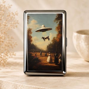 May include: A rectangular silver metal cigarette case featuring a printed scene of a flying saucer hovering over a field with people in period clothing and a cow being abducted by a beam of light in a landscape with trees and a blue sky.