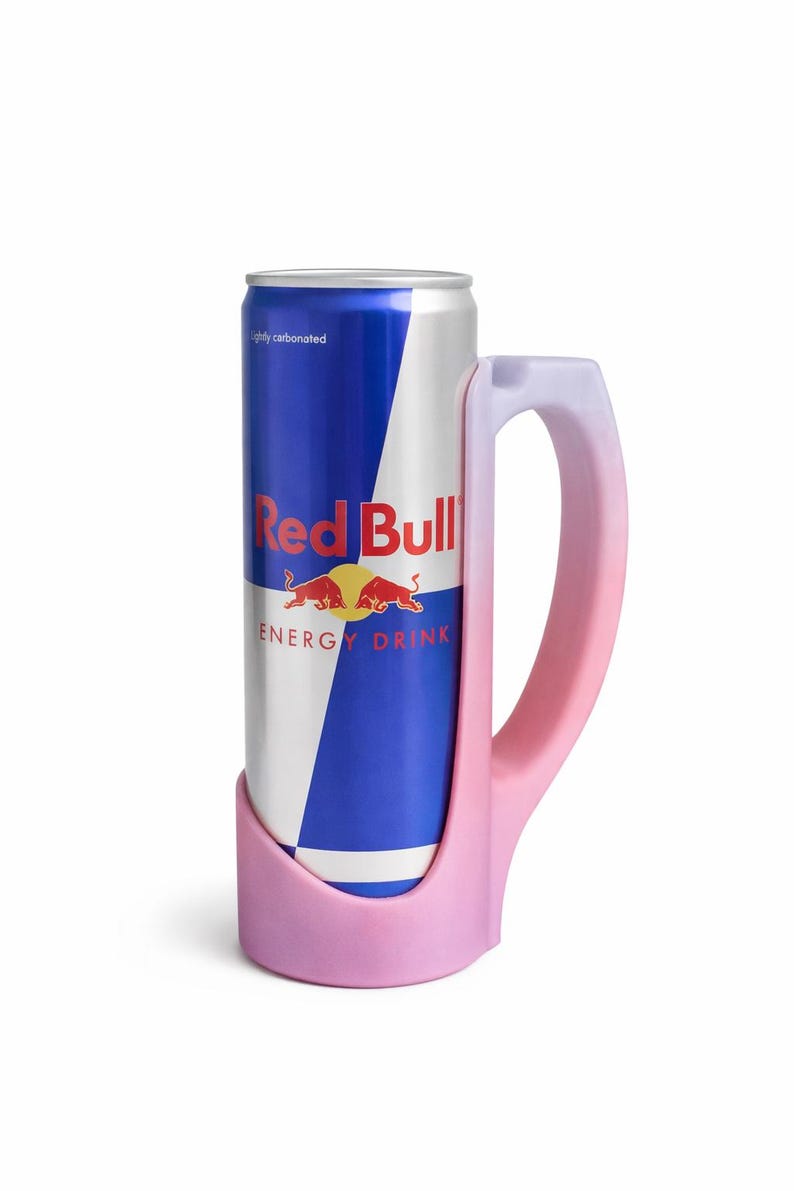 May include: A Red Bull energy drink can held in a pink mug-shaped holder. The can is blue and white with the Red Bull logo. The holder has a handle and is a gradient of pink and white.