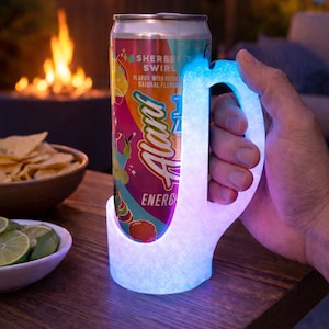 Op de afbeelding: Een gloeiende drankhouder met een handvat, met een blikje "Alani Energy"-drank. Het blikje heeft een kleurrijk ontwerp met de woorden "Sherbet Swirl". De woorden "Glows in the Dark" staan boven de drankhouder.