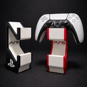 May include: A black and white PS5 controller stand with the PlayStation logo. A white and black stand is on the left, and a red, white, and black stand holds a white and black controller on the right. The PS5 logo is visible.