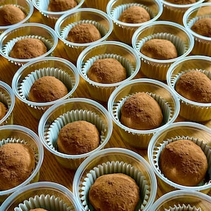 May include: Close-up of numerous chocolate truffles, each dusted with cocoa powder, presented in individual clear plastic cups with white paper liners. The truffles are a rich brown color, and the arrangement suggests a dessert or treat.