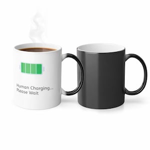 May include: Two ceramic mugs, one white and one black, with black handles. The white mug has a green battery graphic and the text "Human Charging... Please Wait." The white mug contains a dark liquid, with steam rising from it.