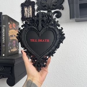 Op de afbeelding: Een zwart hartvormig wandbord met een sierlijk frame in filigraanstijl en een kruis aan de bovenkant. Het midden van het bord is effen zwart met de tekst TILL DEATH in dikke, rode hoofdletters in het middelste gedeelte.