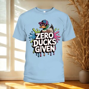 May include: Light blue t-shirt featuring a cartoon duck wearing sunglasses and a cap. The duck is surrounded by colorful paint splatters, with the text "ZERO DUCKS GIVEN" in bold letters.