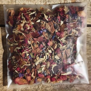 Peut inclure: Un sac en plastique transparent rempli d'un mélange de fleurs et d'herbes séchées. Le contenu est un mélange de teintes rouges, roses, marron et vertes, avec quelques accents bleus et blancs. Le sac est posé sur une surface en bois.