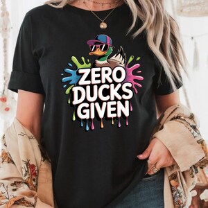 May include: Black t-shirt featuring a cartoon duck wearing sunglasses and a cap, surrounded by colorful paint splatters. The text "ZERO DUCKS GIVEN" is prominently displayed in white, with a playful, casual design.