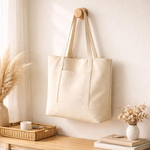 Puede incluir: Un bolso tote de lona color crema con un bolsillo frontal cuelga de una clavija de madera. El bolso tiene asas largas y está suspendido contra una pared de tonos neutros. La escena incluye una bandeja decorativa, un jarrón y libros.