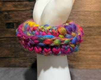 Könnte beinhalten: Ein dickes, grobes Häkelarmband aus einem lebendigen, mehrfarbigen Garnmix in Magenta, Violett, Blau, Gelb und Grün. Das Accessoire weist ein strukturiertes, geschlungenes Maschenmuster auf, das eine dichte, abgerundete Form erzeugt.