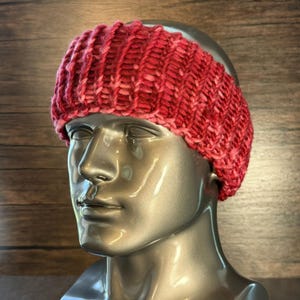 Puede incluir: Una diadema de punto roja y rosa con un diseño acanalado se muestra en una cabeza de maniquí. La diadema está hecha de hilo grueso y tiene un aspecto texturizado. El maniquí es plateado y el fondo es una pared de madera.