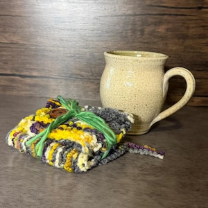 Könnte beinhalten: Ein gesprenkelter beiger Keramikbecher mit geschwungenem Henkel steht neben einem gefalteten, mehrfarbigen Strickquadrat. Der Stoff besteht aus gelbem, violettem und grauem Garn, ist mit einem braunen Holzknopf befestigt und mit grünem Garn verschnürt.