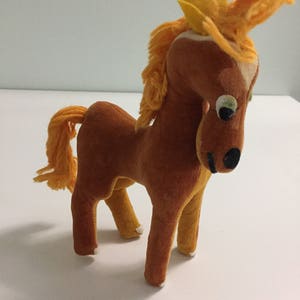 May include: A brown plush horse toy with a yellow mane and tail. The horse has large, black eyes and a black nose. The horse is standing on a white surface.