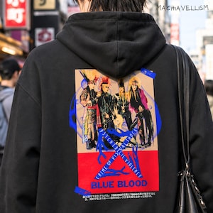 Puede incluir: Una sudadera negra con capucha que presenta un gran gráfico rectangular en la espalda. El estampado muestra una banda con cabello colorido, el texto PSYCHEDELIC VIOLENCE CRIME OF VISUAL SHOCK y BLUE BLOOD, junto con texto japonés.