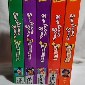 May include: A collection of five Disney "Sing Along Songs" VHS tapes. The spines are green, purple, red, and orange, with titles like "Campout at Walt Disney World" and "Circle of Life". Each tape features Disney characters.