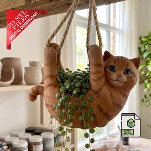 Pode incluir: Um vaso suspenso em forma de gato malhado laranja com olhos azuis, pendurado por duas cordas grossas de juta. A figura do gato tem um centro oco contendo uma planta verde pendente com pequenas folhas redondas. Uma faixa vermelha diz 3D STL FILES.