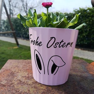 Könnte beinhalten: Ein hellrosa Blumentopf mit dem Text "Frohe Ostern" und Hasenohren. Der Topf enthält eine grüne Pflanze mit einer einzelnen rosa Blüte. Der Topf steht auf einer rostbraunen Oberfläche.