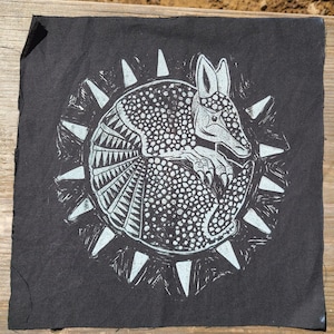 May include: A square black fabric patch featuring a white linocut-style print of a curled armadillo. The armadillo is surrounded by a circular border of radiating triangular spikes, creating a sunburst effect against the dark background.