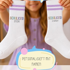 Könnte beinhalten: Ein Paar weiße Socken mit zwei helllila Streifen am oberen Rand. Eine Socke zeigt den Text "SCHULKIND 2026" und die andere "SCHULKIND Leonie" in bunter Schrift. Das Bild enthält Sprechblasen mit dem Text "BEIDE SOCKEN GLEICH ODER 1 NAME + 1 JAHR" und "PERSONALISIERT MIT NAMEN".