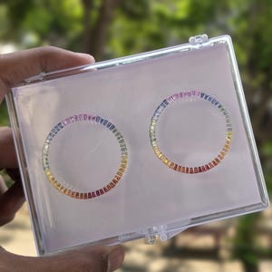 Puede incluir: Dos anillos circulares adornados con un espectro de gemas de colores se presentan dentro de una caja de plástico transparente. Las gemas transitan del rojo al amarillo al azul. La caja se sostiene contra un fondo verde borroso.