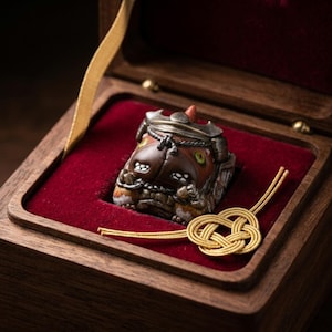 May include: A detailed, artisan-crafted keycap featuring a brown, orange, and bronze design, presented in a wooden box with a red velvet interior. The keycap has intricate details and is accompanied by a gold-colored decorative cord.