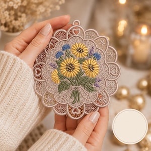 以下が含まれることがあります： 黄色いヒマワリと青いヤグルマギクの花束が刺繍された円形のオーナメント。デザインは、繊細な切り抜き模様が施されたトープ色のスカラップ状のレース風の縁取りで囲まれており、上部には小さなループが付いています。
