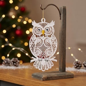 May include: A white lace owl ornament with intricate embroidery and a yellow beak, hanging from a dark brown wooden stand. The owl features detailed swirling patterns, a grid-like chest design, and is perched on a branch, suspended by a gold string.