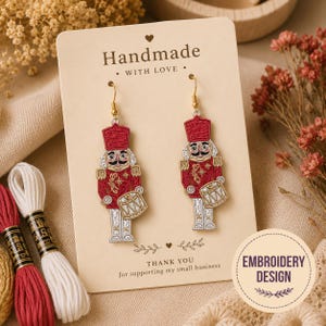 May include: A pair of dangling earrings featuring embroidered nutcracker figures in red, white, and gold thread. The nutcrackers wear tall red hats, red jackets with gold detailing, and hold small drums. The earrings hang from gold-toned hooks.