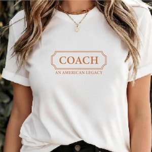 May include: A white short-sleeved t-shirt featuring a rectangular orange graphic on the chest. The graphic contains the word COACH in large capital letters, with the text AN AMERICAN LEGACY printed in smaller capital letters directly underneath.