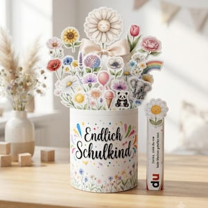 May include: A cylindrical white container with the text Endlich Schulkind and colorful floral illustrations. It holds a bouquet of paper cutouts featuring flowers, a panda, an elephant, and a rainbow. A small rectangular tag with a daisy and text is attached.