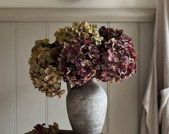 May include: A bouquet of hydrangea flowers in shades of deep burgundy, muted green, and brown, arranged in a weathered, gray, round ceramic jug. The jug sits on a rustic, rectangular wooden stool with a visible grain and textured surface.