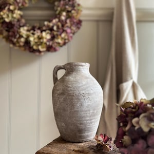 May include: A rustic, light grey ceramic jug with a single side handle and a textured, weathered surface. The vessel features a rounded body and a narrow neck, showcasing a matte finish with subtle horizontal ridges and visible surface imperfections.
