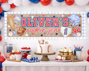 Könnte beinhalten: Eine Baseball-Party-Dekoration mit einem Banner mit der Aufschrift "Oliver's rookie year 1". Das Banner zeigt Baseball-Ausrüstungs-Illustrationen. Ein Kuchen, Cupcakes und Basebälle stehen auf dem Tisch, mit rot-weiß-blauen Luftballons im Hintergrund.