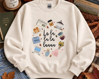 Könnte beinhalten: Ein cremefarbenes Sweatshirt mit Rundhalsausschnitt und einem zentralen Aufdruck mit dem Text La la la la laaaa, umgeben von Illustrationen einer Zeitung, eines Kaffeebechers, eines Pavillons, Pfannkuchen, eines Yale-Pullovers und Blumen.