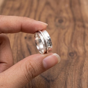 Peut inclure: Une bague en argent avec une bande texturée et un anneau extérieur lisse et rotatif. La bague est tenue sur un fond en bois flou, mettant en valeur sa finition en métal poli. Le design de la bague suggère un accent sur les bijoux et accessoires.